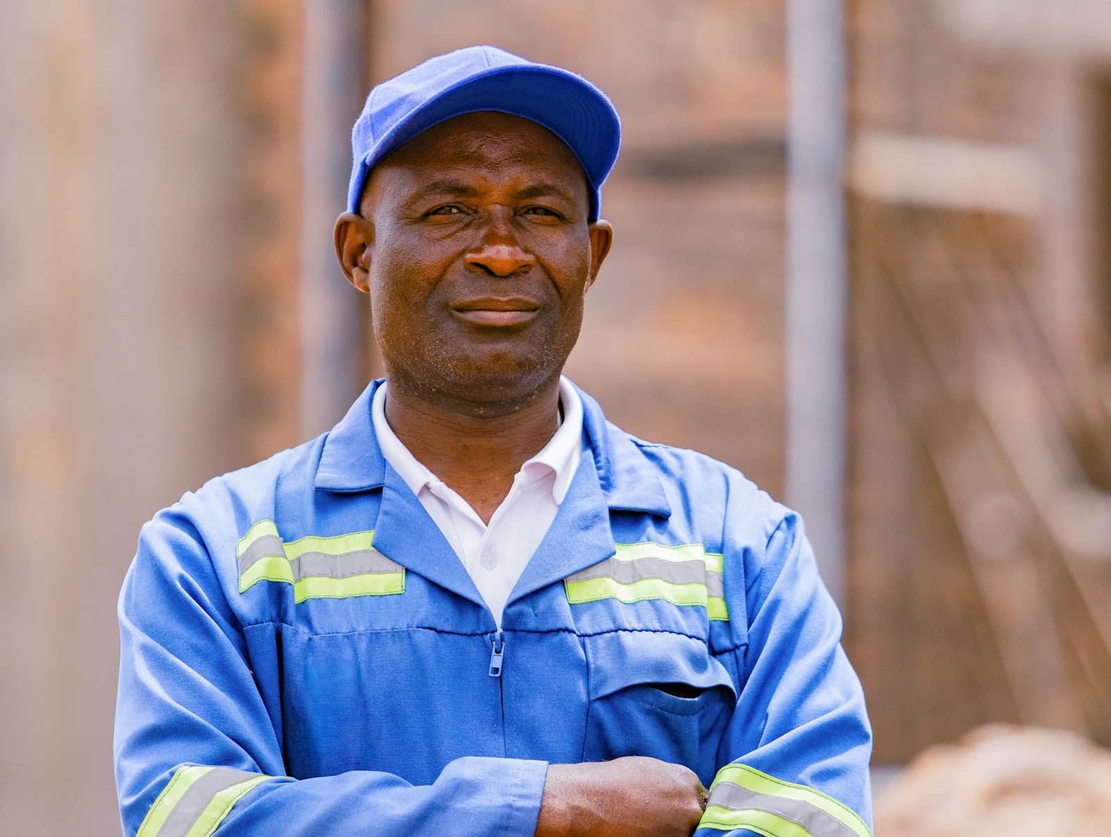 A tradesperson in a blue work uniform and cap, arms crossed, on a job site