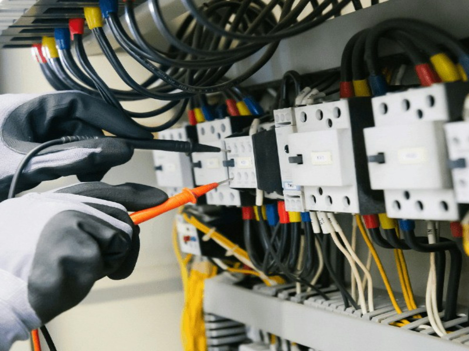A gloved electrician testing a switchboard with multimeter probes among coloured wiring